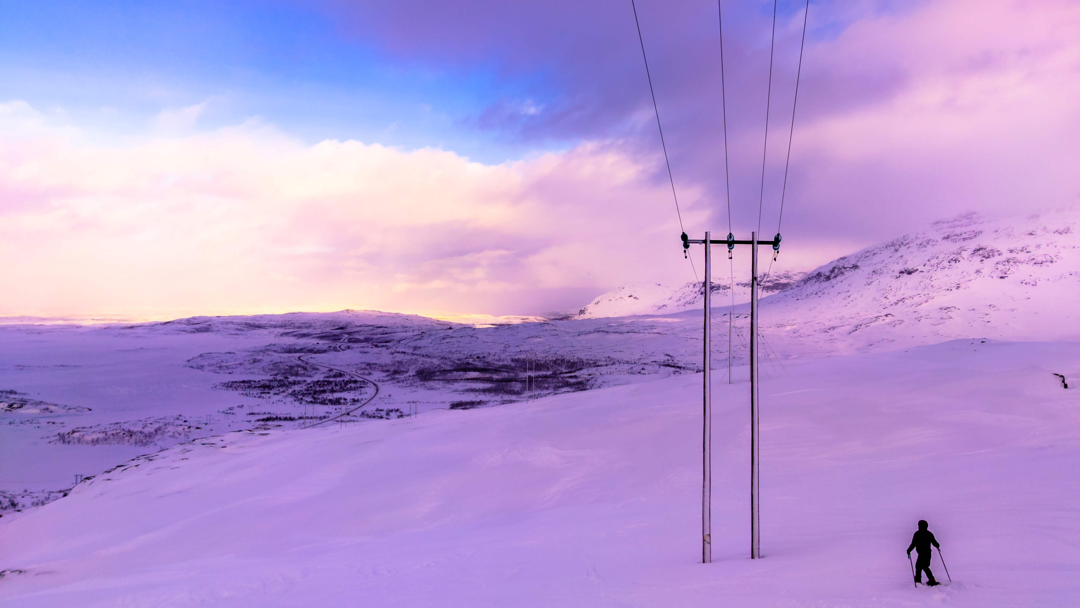 Photo from the top of a snowy mountain with a person skiing towards a powerline, with a road in the background